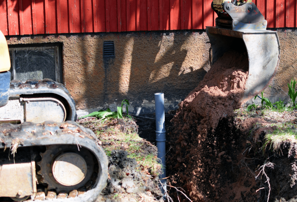 Excavator digging a trench beside a building, with soil being removed around an exposed underground pipe and foundation.