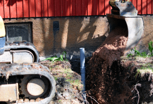 Excavator digging a trench beside a building, with soil being removed around an exposed underground pipe and foundation.
